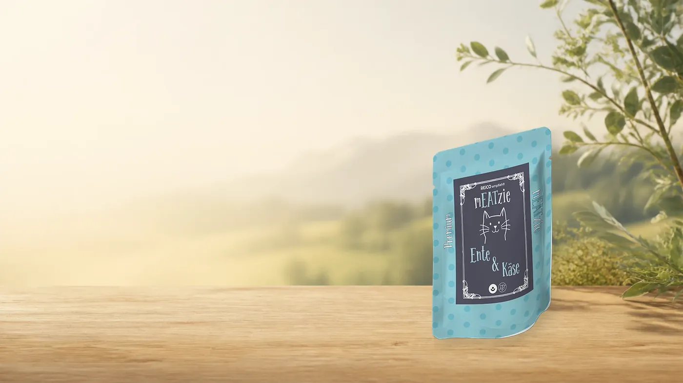mEATzie Ente & Käse – offizielles REiCO Produktmotiv in warmer Naturkulisse mit heller Holzfläche.